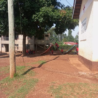 The image depicts an outdoor scene in Kisa Central ward, Kenya, featuring children, buildings, and vegetation under a clear sky. In the foreground, a light-colored wooden utility pole stands on the left, rooted in reddish-brown earth with sparse green grass patches. A dark shadow extends diagonally from the pole across the ground.
In the midground, approximately 10-12 children, predominantly wearing red uniforms, are present. Several children interact with playground equipment, including a red-orange slide with a ladder being climbed by one child, and a light blue-green slide further right. Other children are gathered on the ground beneath a large, mature tree with dense green foliage, located centrally.
Behind the tree, two multi-story concrete buildings are visible. The left building, two stories tall, has a balcony and multiple windows, appearing partially constructed or unfinished. The right building, also concrete and multi-story, shows exposed window frames and an external staircase, similarly suggesting an unfinished state.
On the far right, a cream-colored building with a brown base features a red cross symbol painted on its upper wall and an illegible white informational sign below it. This building has a brown-tiled roof and a concrete strip along its base. The background consists of dense green trees and bushes, with glimpses of red-tiled roofs in the distance, beneath a clear blue sky.