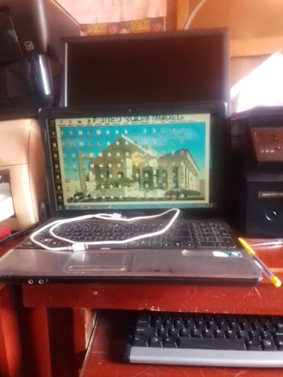 The image shows a laptop computer open on a table in an office in Auchi, Nigeria. The laptop is displaying a computer-generated image of a building. A white cord connects the laptop to a black printer. There is a yellow pen on the table and a black keyboard resting on the lower shelf of the table. The table is brown with a red stain. The laptop screen is showing a computer generated image of a building, and the building is depicted in blue, yellow, and grey. The image was likely taken inside a room because of the lack of natural lighting. The laptop is likely being used to design or view the building. There are no people in the image.