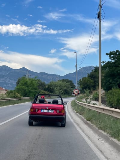 In this roadside scene from Tomba, Montenegro, a red convertible Volkswagen Golf is captured from behind, driving on an asphalt road. The car's license plate reads "BR BA711" with a circular sticker to its right. A person, wearing a red helmet, is visible in the driver's seat, their arm resting on the side of the car. The road is bordered by a metal guardrail on the right, and lush green trees and vegetation line the other side. In the distance, majestic mountains rise against a bright blue sky dotted with fluffy white clouds. A traffic sign indicating a speed limit of 50 is visible on a pole near the roadside. A power line pole with wires stretches across the frame from the left. The image appears to be taken during the day, with clear weather suggesting a pleasant driving experience.