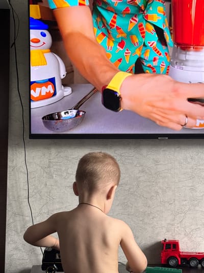 The image shows two subjects: a person's arm and a young child. The arm, adorned with a colorful ice pop patterned shirt and a yellow smartwatch, is positioned over a white toy with a smiling face and blue eyes. The toy also features yellow arms and an orange section with the letters "VM" visible. To the right of the arm, the back of a young child is visible, showing their blonde hair and bare back. The child is leaning towards a toy fire truck, which is red with black wheels and is positioned vertically on a textured, light grey wall. The setting appears to be indoors, with a television screen displaying the arm and toy on the left side of the image. The lighting suggests it might be daytime, though no specific time can be determined. The overall scene appears casual, possibly depicting playtime or a moment of interaction between a child and an adult. The location context is Яромінскі сельскі Савет, Belarus. There is no discernible weather or strong emotional cues.
