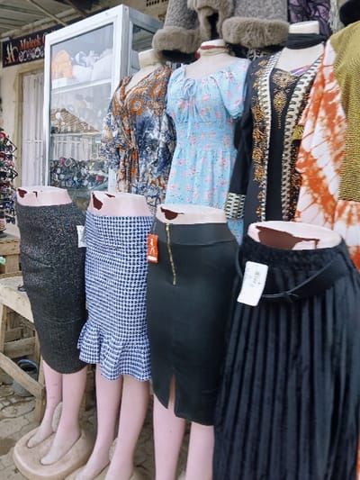 The image displays an outdoor market stall or storefront showcasing clothing on multiple mannequins. The specific location of "Kungulu School, Zambia" is not discernible within the scene.
**Subjects and Objects:**
The foreground features four lower-body mannequins, depicted from the waist down, displaying skirts. From left to right: a dark, textured pencil skirt; a blue and white houndstooth pattern skirt with a ruffled hem; a black pencil skirt with a vertical gold zipper and a front slit; and a black pleated maxi skirt. These mannequins have pale skin tones and flat, bare feet resting on beige circular bases.
Behind these, five upper-body mannequins display tops and dresses. From left to right: a patterned garment in shades of blue and orange/brown; a light blue off-the-shoulder dress with a floral pattern and cinched waist; a black long-sleeved garment with elaborate gold and black embroidery; an orange and white tie-dye pattern garment; and a partial view of a yellow ribbed top. Above these, three additional mannequin heads, partially visible, display fur-trimmed hats.
**Environment:**
Behind the main display, a white-framed glass display case containing various items, possibly sunglasses or small accessories, is visible. To the left of this case, a rack of additional items, possibly more sunglasses, is present. Behind the glass case, a white door or curtain is partially open, and a white sign with "Maleek" in black script is mounted above it. Above the sign, a section of a metal grate or mesh is visible. The ground is paved with interlocking concrete blocks, appearing somewhat dusty. A crude wooden stand and a blue bucket are positioned behind the leftmost leg mannequins. The scene is illuminated by natural light.