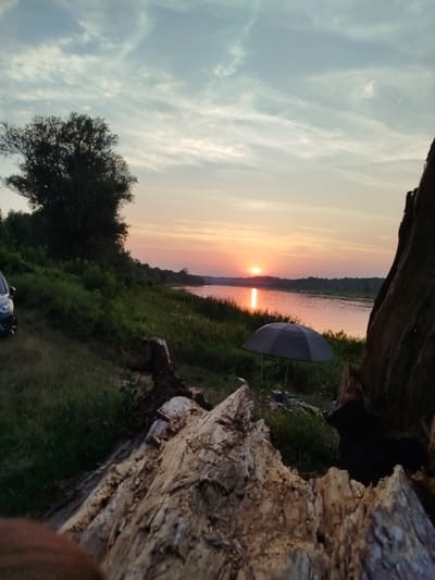 The image depicts a sunset over the Oder River in Topolina, Poland. In the foreground, a large, weathered piece of driftwood occupies the right side of the frame. To the left, a dark-colored car is parked on a grassy embankment, with a dense line of trees and vegetation extending towards the river. A dark grey umbrella is set up on the riverbank, near the water's edge. The river reflects the warm hues of the setting sun, which is visible on the horizon. The sky above is a mix of blue and soft grey clouds. The overall scene is one of tranquility and natural beauty, with elements suggesting a peaceful outdoor setting.