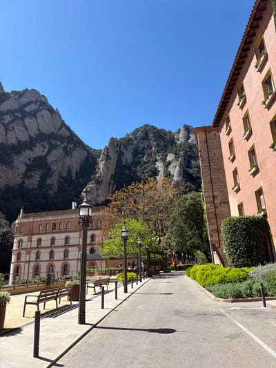 The image captures a sunlit outdoor scene at Monistrol de Montserrat, Spain, featuring a striking blend of natural beauty and architecture. In the background, the iconic, rugged peaks of the Montserrat mountain range dominate, characterized by their unique grey, rocky formations and patches of dark green vegetation, all set against a clear, brilliant blue sky.
Two distinct multi-story buildings frame the scene. On the left, a reddish-orange building with a tiled roof features several windows, each equipped with a planter box containing small green plants. On the right, a lighter-colored building, possibly beige or white, exhibits a series of arched windows with red brick detailing.
A wide, paved pathway or road extends through the center of the image, appearing to be a pedestrian area or courtyard. This path is lined with several dark streetlights and benches. A few indistinct figures can be seen sitting on the benches or walking in the distance, suggesting leisurely activity. Lush green hedges and trees, some with sparse, yellowish-orange leaves, border the path and contribute to the vibrant foreground. Strong shadows cast by the streetlights and buildings indicate bright daylight, suggesting the photograph was taken on a clear, sunny day, likely mid-morning or afternoon. The scene conveys a sense of grandeur and tranquility, with the imposing mountains providing a magnificent backdrop to the well-maintained surroundings. No discernible text is visible in the image.