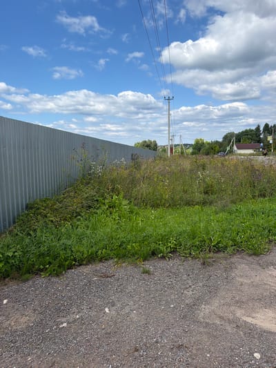 The image depicts an outdoor scene on a partly cloudy day in Stankovo, Russia. A tall, corrugated metal fence runs along the left side of the frame, extending into the distance. Adjacent to the fence is an overgrown area with tall grasses and wildflowers in various shades of yellow, white, and purple. The ground in the foreground is covered with gravel and dirt.
In the mid-ground, a utility pole with power lines is visible. Further in the distance, across a field of wildflowers, a small white house with a dark roof is situated. There are trees lining the horizon, and a few road signs are present. A dark-colored car is parked near the house. A person wearing yellow and black clothing is walking away from the house towards the right side of the frame. Two triangular road signs, possibly indicating a warning or hazard, are placed along the road. The sky is blue with scattered white cumulus clouds.