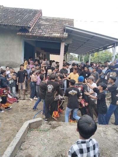 The image depicts an outdoor gathering in Kerjo, Indonesia. A large crowd, estimated at 50-70 individuals, primarily young adults and teenagers, is observing a central activity.
The environment features a single-story building with a brown tiled roof on the left, painted in light blue and beige tones. An attached lean-to structure with a corrugated metal roof extends to the right, sheltering part of the crowd. The ground is uneven, composed of dirt and some concrete, with a concrete curb in the immediate foreground and a blue hose visible on the ground. Trees and foliage are present in the background, behind the lean-to.
In the central area, several individuals are engaged in a performance. At least two performers wear elaborate dark costumes with red and yellow fabric elements, one featuring a decorative headpiece, indicative of a traditional Jaranan (hobby horse) dance. They are surrounded by approximately eight to ten young men who are physically interacting with them, appearing to support, hold, and restrain the performers, whose postures suggest energetic or trance-like movements. One of these men has distinct blue-dyed hair. Several interacting individuals wear black t-shirts, some bearing white text and graphics, including "SATRYA DHARMA" and "SINGA NDOWO".
The surrounding crowd is densely packed, standing and watching the central event. A child, seen from behind wearing a black and white plaid shirt, is in the foreground, facing the activity.