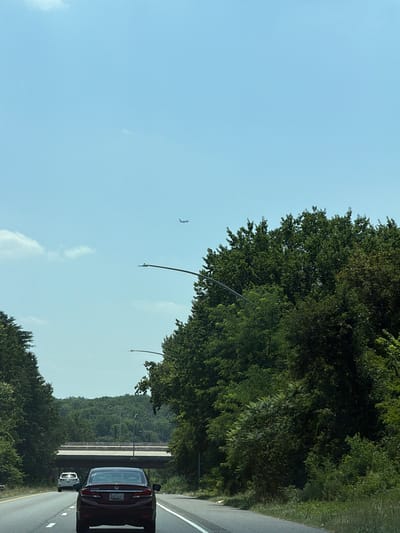 The image depicts a view from inside a moving vehicle on a highway in Glen Burnie, Maryland, United States of America. The sky is clear and blue with a few white clouds. In the distance, a small airplane can be seen flying. The highway is lined with lush green trees on both sides. A concrete overpass is visible in the background, with a few cars on the road beneath it. In the foreground, the rear of a dark-colored sedan is the primary subject, with its license plate clearly visible. Another white car is visible further ahead on the same lane. Streetlights with their lamps are intermittently placed along the side of the highway. The scene suggests a daytime journey on a sunny day.