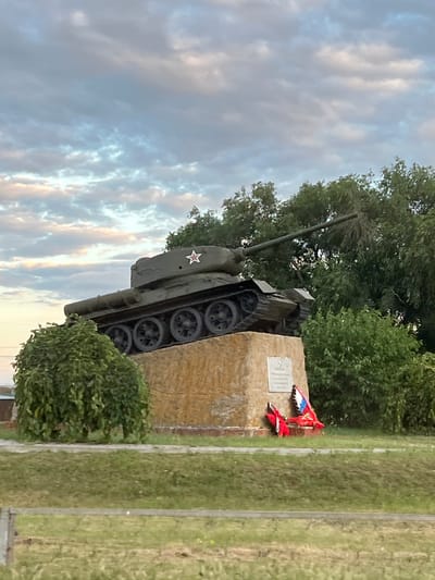 The image shows a Soviet-era T-34 tank monument at the entrance to the city of Zimovniki, Russia. The tank, painted olive drab, is mounted on a concrete pedestal covered with light-colored stone. A red star with a white outline is visible on the side of the tank's turret. In front of the pedestal, there is a plaque and a memorial wreath with red and blue ribbons. The monument is surrounded by green trees and bushes, with a grassy area in the foreground. The sky above is cloudy, with hues of gray and pale yellow, suggesting either dawn or dusk.