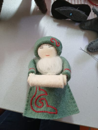 The image shows a close-up of a felt doll lying on a light-colored, textured surface, likely a table. The doll is green with red decorative stitching on its dress and hood. It has black embroidered eyes and a small red mouth. The doll's arms are crossed in front, holding a rolled-up piece of white felt. Other craft materials, including dark fabric shapes and thread, are visible in the blurred background. The scene does not provide any visual cues that would allow for identification of a specific location in Taraz, Kazakhstan.