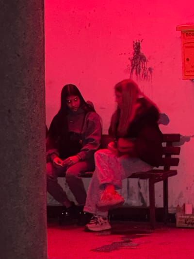 Two people are observed in a red-lit environment. One individual, facing the camera, is wearing a dark hooded jacket. The other, with long blonde hair, is leaning over a bench. A wall with a sign and some markings is visible in the background. The image was taken from a low angle. It is unknown if the scene is located in the city of Mihailovo, Bulgaria.