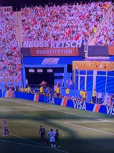 The image depicts a crowd of spectators watching a sporting event from a stadium. A large electronic billboard displays the text "NEUSS KETSCH SUBSTITUTION". The stadium appears to be modern, with blue and orange accents on the seating area. In the foreground, several athletes in uniform are visible on a green field. One athlete, wearing a white jersey with the number 2, is facing away from the camera. Another athlete, dressed in a dark jersey, stands to his left. A group of athletes in striped jerseys is also visible in the distance. The stadium is likely hosting a soccer match. This scene does not provide any information regarding the city of Bahovitsa, Bulgaria.
