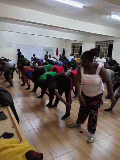 An indoor scene, likely a community hall or exercise studio in Riara Ridge, Kenya, depicts a large group of adults engaged in a group physical activity. The room features light-colored walls, a light wooden tiled floor, and ceiling-mounted fluorescent lights. On the left wall, a whiteboard with some faint, illegible markings is visible, while two windows with grilles on the right side appear dark outside, suggesting the event is taking place in the evening or at night.
Most participants are actively performing a movement, bent forward at the waist with their hands reaching towards their feet or the floor, indicative of stretching, exercise, or a dance routine. They are dressed in casual and comfortable attire, including t-shirts, track pants, and a mix of athletic shoes and casual footwear. In the immediate foreground on the right, a woman wears a white tank top, patterned trousers, and light green flat shoes. Another individual in the mid-ground left is dressed in a black top and black and red striped pants, also in a bent posture.
The atmosphere suggests focused participation, with individuals concentrating on the collective movements. A wooden table or bench draped with yellow fabric is partially visible in the foreground on the left. No clear, legible text is visible within the image.