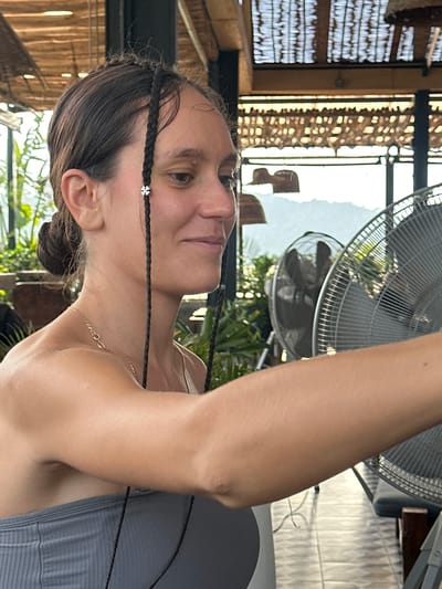 This image features a woman in a semi-outdoor, bright setting, likely in Hà Giang 1 Ward, Vietnam. The woman is captured in a close-up, chest-up shot, her head tilted slightly upwards and to her right, with a calm expression. She has dark hair, styled with several thin braids, one of which is adorned with a small, silver, clover-shaped charm near her temple. She wears a delicate gold chain necklace and a grey ribbed top. Her right arm is raised and partially visible towards the upper right of the frame.
The background suggests a tropical or rustic architectural style. To the left, there are slatted wooden or bamboo walls, creating patterns of light and shadow from the bright daylight streaming in. On the right, two large metal-grille standing fans are visible, indicating warm weather. Green foliage and plants are seen through openings in the structure, further enhancing the outdoor or open-air ambiance. The overall scene conveys a relaxed atmosphere in a sunny location. No visible text is present in the image.