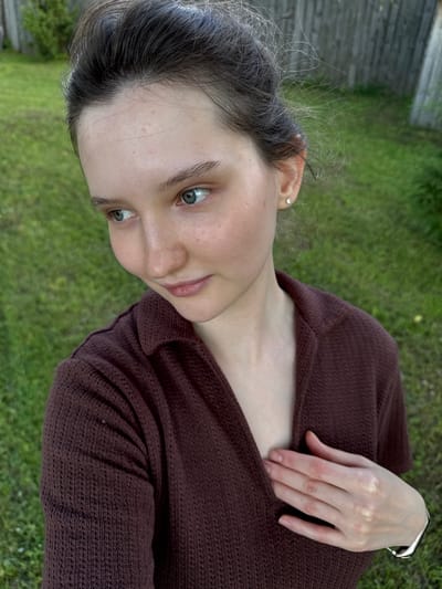 In Burdyata, Russia, a young woman with light skin, light brown hair, and blue eyes is pictured outdoors. She is wearing a dark brown, collared, knitted shirt. Her hair is pulled up loosely, with some strands loose around her face and ears. She has a small, square stud earring in her right ear. Her left hand is resting on her chest, near her collarbone, and she is wearing a silver bracelet on her left wrist. The background consists of green grass and a wooden fence. The image appears to be a selfie, taken from a slightly elevated angle, with the woman looking to her left.