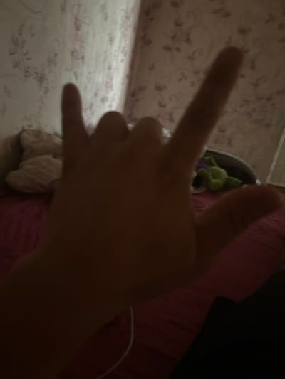 In a dimly lit room, a person's hand is raised in the "rock on" gesture, with the index finger and pinky finger extended, and the thumb, middle, and ring fingers curled down. The hand is positioned in the foreground, obscuring much of the background. Behind the hand, there appears to be a bed with a pink or red quilted comforter. To the left of the bed, several stuffed objects, possibly pillows, are visible. Further back, on the wall, a floral wallpaper pattern can be discerned. In the mid-ground, to the right of the hand, a gray bin or basket contains what appears to be a green plush toy. The overall scene suggests an indoor setting, possibly a bedroom. There is no information available to indicate the location in the city of Saran, Kazakhstan.