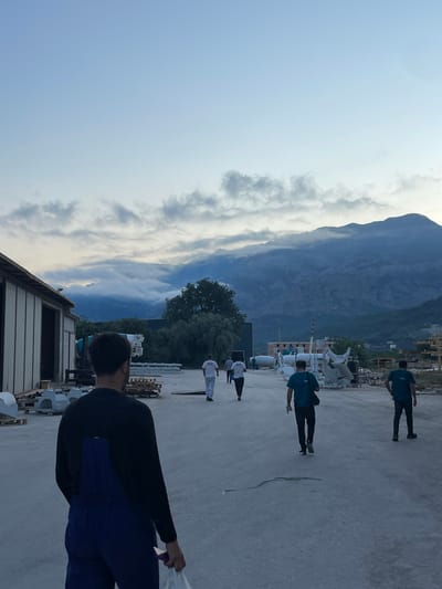 The image captures an outdoor scene at what appears to be an industrial or construction site in Tomba, Montenegro. The sky is a pale blue with scattered, low-lying clouds that are beginning to obscure the tops of distant, rugged mountains. In the foreground, a man with dark hair is seen from the back, wearing a black long-sleeved shirt and blue overalls. He is holding a white plastic bag in his right hand. Several other individuals are walking away from the camera across a paved area. Some are wearing dark pants and teal shirts, while others are in white t-shirts and light-colored pants. Various pieces of industrial equipment, possibly cement mixers or large pre-fabricated components, are visible throughout the scene. Pallets of materials are stacked on the ground. A long, low-slung building with white siding is situated to the left of the frame, with its entrance open. Trees and more construction materials are visible in the middle ground and background. The overall lighting suggests it is late afternoon or early evening.