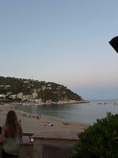 The image depicts a serene coastal scene in Llafranc, Spain, during what appears to be late afternoon or early evening. The sky is a gradient of pale blue to soft pink. In the foreground, a woman with long blonde hair is seen from behind, wearing a light-colored strapless top and black shorts. She is holding a patterned bag. To her left, a metal bench is visible.
The middle ground features a sandy beach populated by several groups of people. Some are sitting or lying on towels, others are standing or walking. Children are present, and one person appears to be kicking a soccer ball. The beach is bordered by the calm, grey water of the Mediterranean Sea.
Dominating the background is a lush, green hillside covered in trees and dotted with buildings, including houses and a harbor filled with numerous white boats. A construction crane is visible on the hillside. The coastline curves into the distance, with more buildings and trees along the shore. The overall atmosphere is one of peaceful relaxation and summer leisure.