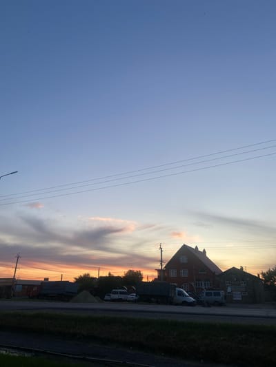 This is a daytime, medium-shot, eye-level, exterior photograph of a street in Egorlykskaya, Russia, at sunset. The sky is a gradient of blue to orange and pink as the sun sets. Power lines are visible across the sky, as well as a street lamp on the left. In the middle ground, there are several buildings, including a large red house with multiple windows and a smaller stone building with a sign. Several vehicles, including a truck and a van, are parked on the street in front of the buildings. The foreground shows a strip of grass and a ditch.