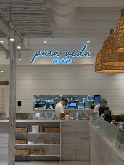 This indoor shot shows the interior of Pura Vida in Coral Gables, United States of America. The neon sign for Pura Vida is on the wall. Below the sign are wooden decorative beams. A worker in a white shirt is standing at the counter, seemingly using a phone. Another person is behind the counter, wearing a white shirt and cap, and working on a computer. Near the workers, there is a storage shelf with wicker baskets and other objects. A wicker decoration is at the top of the shot.