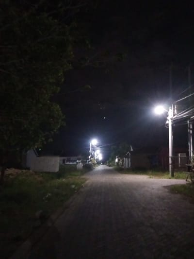 This media file depicts a dimly lit, paved street at night in Tibojong, Indonesia. The scene is illuminated by streetlights, creating a moody and quiet atmosphere. On the left side of the street, a large tree with dark foliage casts shadows, and a simple, light-colored building is partially visible. The right side of the street features more buildings, some with visible windows, and more streetlights. The paved road is textured and appears to be a pathway or residential street. The sky is dark, suggesting it is nighttime. There is no visible text in the image. The overall impression is one of a peaceful, albeit somewhat stark, nighttime setting in a residential area.