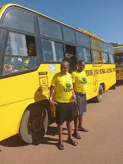 The image depicts an outdoor scene in Shirungu-Mugai ward, Kenya, under a clear blue sky. Two adult women stand in the foreground on a light brown, unpaved surface, facing the viewer. Both wear yellow short-sleeved t-shirts bearing a green circular "UDA KAZI NI KAZI" logo, dark-colored skirts, and open-toed sandals. The woman on the left holds a dark smartphone in her left hand. Behind them and to their left, a yellow school bus is parked. The bus's side displays "FRIENDS HIGH SCHOOL - SILUNGAI" in black lettering and a yellow sign with "SILUNGAI" and a crest. A person, also in a yellow UDA t-shirt, is visible through the bus's front passenger-side window, looking outwards. A yellow ladder is affixed to the side of the bus. A second yellow bus is partially visible in the background to the right. Shadows are cast by the individuals and the bus, indicating direct sunlight.