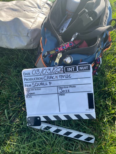 The image depicts an outdoor filmmaking scene in Chonas-l'Amballan, France, under natural daylight conditions. The foreground is dominated by a white film clapperboard with a hinged black and white striped stick, lying open on lush green grass containing clover. The clapperboard displays handwritten details: "DATE 03/03/226", "PRODUCTION CRACK MUSIC", "FILM SQUALL P", "Mise en Scène Joan", and "Directeur Photo Joji". The "INT" (Interior) indicator is marked, alongside "NUIT" (Night), though the setting appears to be exterior and daytime. A small "PANASTORE" logo is present on the lower right of the clapperboard's white surface.
Behind and to the left of the clapperboard is a partially open blue and orange backpack. Its contents are visible, including a white spray can, various papers or cards, and a red object. Attached to the backpack's exterior straps are multiple carabiner clips, one holding a colorful woven lanyard with a "SALEWA" tag. A prominent gold-colored zipper pull hangs from a pocket. A metallic pin or badge depicting a cartoonish character, possibly a skull or alien head, is also visible. To the left and slightly behind the backpack, a light grey, possibly fabric, object is partially discernible on the grass. No individuals are present in the frame.
