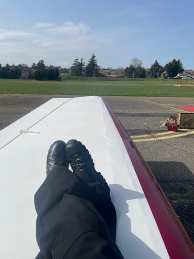 The image depicts a first-person perspective, looking over a person's lower legs and feet resting on the wing of a small aircraft at an aerodrome in Chonas-l'Amballan, France. The individual wears black trousers and black high-top boots with textured soles and metal lacing hardware. Their feet are positioned on the white upper surface of the aircraft wing, which features a prominent red stripe along its edge. A small, white vent-like object is visible on the wing near the left boot.
To the right of the aircraft wing, an asphalt taxiway or runway with yellow painted markings is visible. Further down the taxiway, two objects are present: a red rectangular container or box, and a lighter-colored, possibly wooden, rectangular crate or piece of ground equipment.
The midground consists of a large expanse of manicured green grass, characteristic of an airfield. The background features a rural landscape under a clear blue sky with sparse clouds. A line of trees, including both evergreens and bare deciduous trees, runs along the horizon. Behind the trees, several residential buildings with terracotta roofs are discernible, along with utility poles. The scene is illuminated by natural daylight, casting shadows to the right, suggesting the sun is positioned to the left.