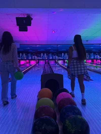 Two young women are bowling in a dimly lit bowling alley. The alley is illuminated by purple and blue lights, casting a neon glow on the lanes and the surrounding area. The woman on the left is wearing a light-colored sweater and jeans, holding a green bowling ball. The woman on the right is wearing a white tank top and a plaid shirt, with her back to the camera. A row of colorful bowling balls is lined up in the foreground, including orange, yellow, red, pink, blue, and green balls. The bowling lanes extend into the distance, with pins visible at the end of each lane. The scene appears to be a casual outing, with the two women engaged in a game of bowling. The location is stated to be Waynesboro, United States of America, but there are no specific visual cues in the image to confirm this precise location.