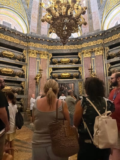 The image depicts the Royal Pantheon within the Monastery of San Lorenzo de El Escorial, located in San Lorenzo de El Escorial, Spain. A group of people are observing the ornate mausoleums lining the circular walls of the pantheon. The architecture is characterized by rich gold detailing against dark marble and pink columns. A large, elaborate chandelier hangs from the domed ceiling. Several individuals are visible from behind, some carrying bags and backpacks. The scene is one of respectful observation within a historically significant burial site.