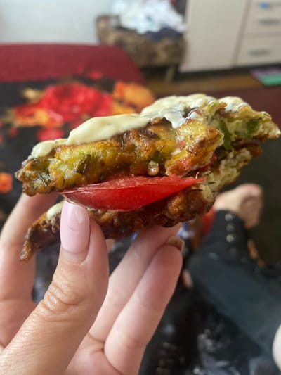 A close-up shot shows a person holding a piece of zucchini fritter with a slice of tomato and a dollop of mayonnaise. The fritter appears to be seasoned with herbs. The background is blurred, but a floral pattern on what appears to be a bedspread or fabric is visible, along with a chest of drawers. The scene is set indoors, likely in a residential environment. The person's hand has well-maintained fingernails with a light pink polish. The bottom right of the image shows a portion of another person's legs and feet, clad in dark clothing. The location of Kisliakovskaya, Russia is not discernible from the visual information in the image.
