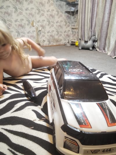 The image shows a young child lying on a zebra-striped blanket in a room. The child is partially visible, with blonde hair and appearing to be naked from the waist up. In front of the child is a white toy SUV with black accents and red and orange racing stripes. The car has a Toyota logo on the roof and text on the front that reads "VSP 444". To the left of the car is a black, elongated object, possibly a toy or electronic device. In the background, there is wallpaper with a floral pattern, a grey inflatable horse toy, and a yellow ball. There are also curtains in the background. The setting appears to be indoors, likely a living room or playroom, with carpeting on the floor. The lighting suggests it could be daytime.