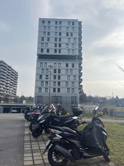 The image captures an urban or suburban scene, likely in Meyrin, Switzerland, under an overcast daytime sky. In the foreground and midground, a row of several black scooters or motorcycles is parked on a paved surface, which consists of gray interlocking bricks on the right and asphalt further left. One prominent scooter displays the text "TET" on its side, and a black helmet rests on the seat of another. A utility pole is visible to the left of the parked vehicles.

In the background, two distinctive modern buildings dominate the landscape. The closer and taller building is a multi-story high-rise with a light gray facade, featuring a grid pattern of rectangular windows and a unique, jagged or sawtooth-like roofline. Further behind and to the left, a second, shorter building exhibits a darker, undulating or wavy facade. Between the parking area and the buildings, a strip of sparse grass and some bare trees are visible, suggesting a season outside of full foliage. No people are present in the scene. The overall atmosphere is quiet and functional, typical of an urban parking area.