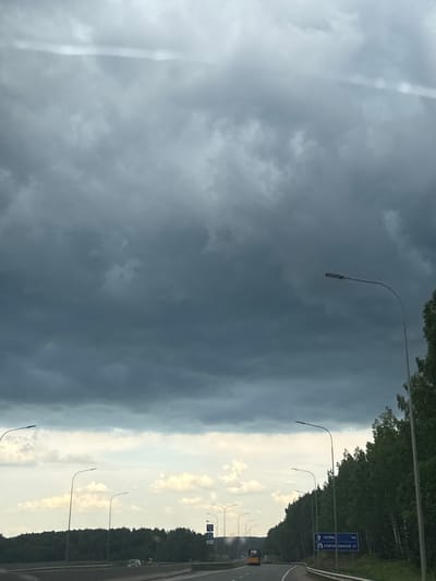 This is an outdoor, eye-level, medium shot captured on a cloudy day. The scene depicts a highway in Beloborodovo, Russia, with a focus on the road, surrounding vegetation, and the sky. The highway appears to be multi-lane, with concrete barriers separating the lanes. Several streetlights line the side of the road, their modern, curved designs contrasting with the natural elements.

On the right side of the highway, a dense forest of birch and other deciduous trees creates a vibrant green backdrop. The trees are tall and appear healthy, with sunlight filtering through the canopy in places, suggesting it is daytime.

The sky is dominated by heavy, dark gray clouds, indicative of an approaching storm or overcast weather. A thin, bright horizontal line is visible in the upper portion of the sky, possibly a contrail or a reflection. In the distance, a few lighter, scattered clouds can be seen near the horizon.

Road signs are visible, including a blue sign with white text and directional arrows, indicating destinations and distances. One sign displays "ПЕРМЬ" (Perm) with the number "64" next to it, suggesting a distance of 64 kilometers to Perm. Another sign beneath it indicates "ГРИГОРЬЕВСКАЯ 37" (Grigoryevskaya 37), likely a street or location name with a distance of 37.

A yellow bus is visible on the highway, traveling in the same direction as the camera. Further down the road, a silver car is seen, also moving along the highway. There are no visible people in the frame, emphasizing the vastness of the landscape and the journey. The overall atmosphere is one of travel under potentially dramatic weather conditions.