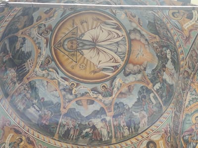 The image shows a detailed fresco painting on the interior dome of the Rila Monastery in Rila, Bulgaria. The central element of the fresco depicts God the Father, rendered as an elderly man with a long grey beard, enclosed within a triangle that symbolizes the Holy Trinity. He is surrounded by a halo and is positioned within a circular frame, with rays of light emanating from him.
The fresco extends to other sections, featuring various biblical scenes and figures. To the left of the central depiction, there is a scene with figures and architecture, possibly representing a biblical event or a historical narrative. Further down, another circular panel contains figures and a landscape.
To the right of the central God the Father, a dove, representing the Holy Spirit, descends amidst clouds and light. Below this, there are arched sections containing painted figures of angels and humans, interspersed with scrolling banners bearing inscriptions in Cyrillic script. These figures are engaged in various actions, some appearing to be in conversation or contemplation.
The overall style of the fresco is religious and symbolic, characteristic of Orthodox Christian art. The colors are rich and varied, with blues, greens, yellows, and reds dominating the palette. The painting's intricate details and vibrant imagery suggest a significant artistic and spiritual undertaking. The Rila Monastery, a UNESCO World Heritage site, is renowned for its well-preserved frescoes and its historical importance.