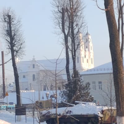 A snowy winter scene captures a partial view of a tan and green military tank, partly covered in snow. Bare trees frame a white church or cathedral with distinctive towers in the background. A building with windows and a white roofline sits between the church and the tank, blending into the snow-covered landscape. The scene is bright, with the winter sun casting shadows on the snow.