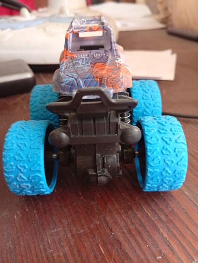 A close-up, overhead shot captures a toy monster truck with bright blue tires, black suspension, and a colorful body featuring orange and blue patterns. The truck is positioned on a dark wooden surface, possibly a table, with some scattered dust visible. In the blurred background, other objects, including what appears to be papers and a white electronic device, are faintly discernible. The image lacks any specific identifying features that would allow for its localization to Kyshtym, Russia, or any other geographical location. The scene is static and devoid of any human presence or activity.