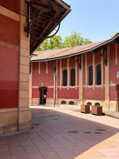 The image depicts the exterior of a building with a red facade and a tiled roof. The building has several arched windows and is constructed with stone elements at the corners and around the windows. In the foreground, a paved courtyard is visible, with square tiles in a reddish-brown hue. Two large, square, rusted metal planters are placed in the courtyard. A shadowed area falls across the courtyard from an unseen structure. The scene appears to be an outdoor public space, possibly part of a historical or cultural institution. The location is identified as the Biblioteca Publica Josep Coll i Vehí in Palma, Spain. There are no people visible in the image.