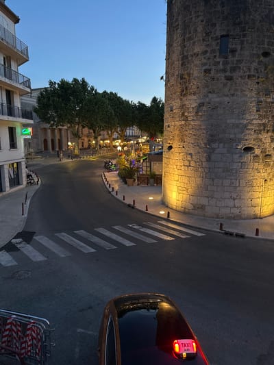The image captures a street scene in the city of Anduze, France, at dusk. A large, ancient stone tower dominates the right side of the frame, illuminated by warm, yellow lights at its base. To the left, a multi-story building with balconies and a green bank sign is visible. A pedestrian crossing with white stripes is painted on the dark asphalt road, which curves through the scene. Several trees with lush green foliage are planted along the street, with lampposts casting a glow on the pavement. In the foreground, the roof of a brown taxi with a lit "TAXI ALES" sign is visible. A metal barricade with red and white striped tape is positioned on the left side of the road. A few motorcycles are parked near a row of outdoor seating and potted plants in the middle ground. Two individuals are walking in the distance on the left side of the street, near a building with columns. The sky above is a deep blue, indicating twilight.
