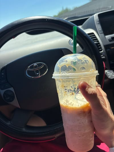 The image shows a close-up of a person's hand holding a clear plastic cup with a green straw. The cup contains a blended beverage with a caramel drizzle. The word "Enjoy" is written in black marker on the side of the cup. The cup is positioned in front of a car's steering wheel, which has the Toyota logo in the center. The car appears to be parked, as indicated by the bright daylight and clear blue sky visible through the windshield. The scene is set inside a car, likely in a suburban or urban environment, given the presence of other vehicles and infrastructure in the background, suggesting it could be in Rancho Cordova, California.