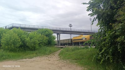 In the city of Luza, Russia, a dirt path leads through a grassy area towards a railway line. An elevated pedestrian bridge crosses over the tracks. On the tracks, a train is visible, including several freight cars and a yellow tank car labeled "GAZPROM" in Cyrillic. The sky is overcast, and trees and foliage are present on both sides of the scene. A person can be seen walking on the pedestrian bridge. The timestamp "2026/06/28 18:08" is present in the lower left corner of the image.