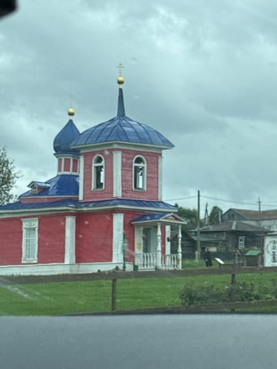 The image shows a brightly colored red and blue wooden church with white trim, located in Nizhnyaya Sinyachikha, Russia. The church features a distinctive blue multi-tiered roof topped with golden crosses. The main body of the church is painted red and has white pilasters and arched windows. There are multiple domes, with the largest dome a vibrant blue and topped with a golden cross. To the left of the church, a small part of a tree with green foliage is visible. In the background, several other wooden buildings, likely houses or other structures, are present, some with dark roofs. A wooden fence runs across the foreground, separating a grassy area from what appears to be a road or paved surface. The sky is overcast with clouds. A person in dark clothing is partially visible in the middle ground to the right of the church, near a small green shed.