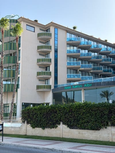 This is a vertical-format photograph of the Hotel Kaktus Albir in l'Alfàs del Pi, Spain. The hotel building is a multi-story structure with a distinctive triangular facade on its upper floors, featuring numerous balconies with green accents and triangular window frames. The lower floors have a more conventional design with rectangular windows.

The building is clad in a light-colored brick or stucco. A large, dense hedge runs along the front of the hotel, obscuring the ground floor in some areas. A paved walkway with a geometric pattern runs alongside the hedge and the hotel. Palm trees are visible in the foreground, with their fronds partially obscuring the lower levels of the hotel.

A sign on the glass facade of the hotel reads "Hotel Kaktus Albir" in green, vertically oriented text. The scene appears to be set during the daytime, with clear blue skies suggesting fair weather. The overall impression is one of a sunny day at a coastal hotel. There are no people clearly visible in the image, and no specific activity is taking place. The architecture of the hotel is the primary focus, highlighting its unique design features.