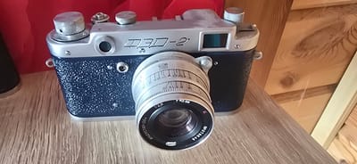 This image displays a vintage Zenit-3M film camera, which appears to be in a state of moderate use. The camera is dark blue with a silver top plate and a silver lens. The lens has markings indicating an aperture of f=2.8 and the serial number "№ 078646". The camera is positioned on a light-colored wooden surface, which may be a shelf or table. In the background, a red fabric and unfinished wooden planks are visible, suggesting an indoor setting. The overall impression is one of a personal collection or display of a historical photographic artifact. There is no information available about the specific location within Gostagayevskaya, Russia, other than it is a city in Russia.