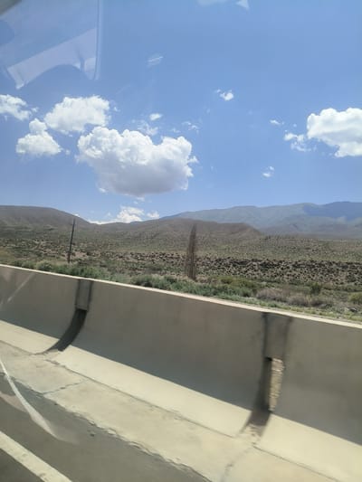 The image captures a view from a vehicle, looking out at a hilly, arid landscape under a partly cloudy blue sky. In the foreground, a concrete barrier runs along the side of the road, casting shadows. Beyond the barrier, the terrain is covered with sparse vegetation and scrub. In the middle ground, a single, tall, thin tree or pole stands out against the hillside. Further in the distance, layered mountains recede into the horizon. The sky is a vibrant blue with scattered white clouds, one large cloud dominating the upper left portion of the frame. There are no people or animals visible in the scene. The overall impression is one of a dry, undeveloped natural environment. This scene is likely located in or around Kok Moynok Eki, Kyrgyzstan, given the mountainous and arid terrain typical of the region.