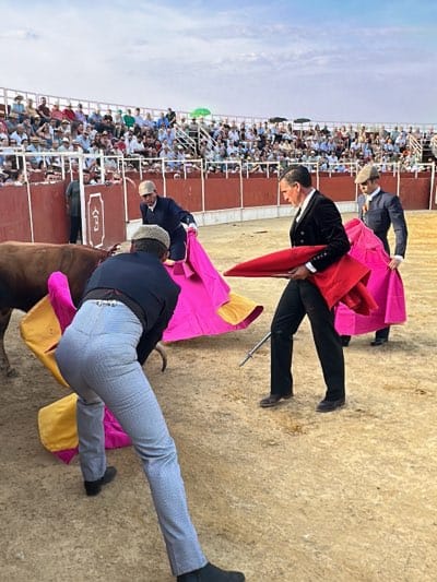 In a bullring in San José del Valle, Spain, multiple bullfighters are engaged in a bullfight. They are dressed in traditional attire, including dark suits, hats, and brightly colored capes. One man, in the foreground, is wearing checkered pants. The bullfighters interact with a bull, which is partially visible. A crowd of spectators is seated in the background, observing the event. The bullring is enclosed by a wall. The ground is dirt or sand. The sky above is partly cloudy.