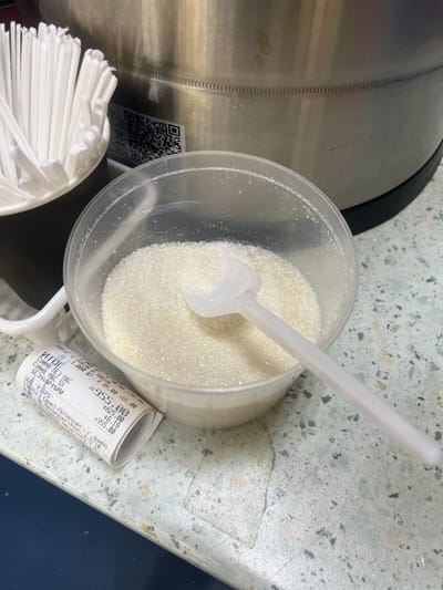 This media file shows a close-up, overhead view of a clear plastic container filled with granulated sugar, resting on a speckled countertop. A white plastic scoop is partially submerged in the sugar. To the right of the sugar container, a rolled-up receipt is placed on the counter. In the lower left corner, a stack of white plastic cutlery is visible. To the left of the sugar container, a portion of a large, metallic cylinder with a QR code sticker is visible.

The receipt shows text in Russian, indicating a purchase from "000 'РЭТРО'" in "Санкт-Петербург" (Saint Petersburg). It details items like "ПИРОГ С луком и яйцом" (pie with onion and egg) and various price calculations. The total amount shown is "955.00".

The scene appears to be in a food service setting, possibly a cafe or a buffet, given the presence of sugar, cutlery, and a receipt from a food vendor. The lighting suggests an indoor environment, likely during the day. The overall mood is neutral and observational, focusing on everyday objects.
