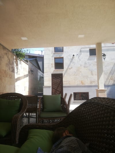 A person, likely a child, is seen sleeping on a couch under an awning in a courtyard. The couch is made of wicker and has green cushions. The background shows a building with stone walls, windows, and decorative metalwork. The scene appears to be in Akhalkalaki, Georgia.