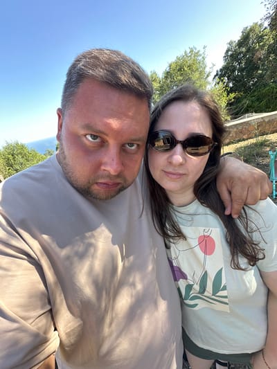 In a selfie taken outdoors, a man and a woman stand close together, with the man's arm around the woman's shoulder. They are positioned in what appears to be a Mediterranean setting, with lush greenery and a distant view of the sea. The man, with a beard and short hair, wears a light-colored t-shirt and looks directly at the camera with a serious expression. The woman, with long brown hair and sunglasses, smiles subtly. The background features trees, a stone wall, and what looks like a patio or garden area. The lighting suggests it is a sunny day. The location is identified as Santa Cristina d'Aro, Spain, a coastal town known for its beaches and natural beauty.