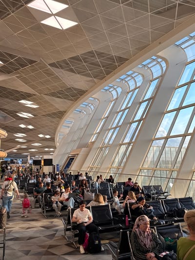 The image depicts a busy airport waiting area, bathed in natural light filtering through a large, arched, glass ceiling. The architecture is modern and spacious, with a geometric tiled ceiling and a long, curved section of windows. Numerous people are seated in rows of dark chairs, facing various directions, suggesting they are waiting for flights. Some individuals are looking at their phones, while others are engaged in conversation or resting. The atmosphere appears to be one of relaxed anticipation, typical of an airport environment. There are suitcases and bags scattered around the seating area. The sunlight streaming through the windows indicates it is likely daytime. The geometric patterns on the ceiling and floor, along with the modern seating, are consistent with a contemporary airport terminal. While specific text is not clearly legible, there are signs visible in the background which are common in airport terminals.