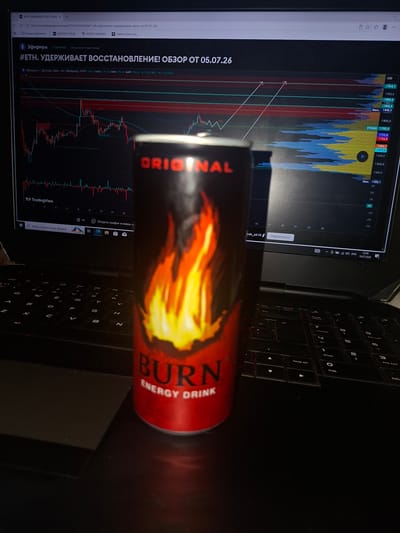 A red and black can of "BURN ENERGY DRINK" is positioned in the foreground, slightly to the right of the center of the frame. Behind the can, a laptop screen displays a cryptocurrency trading chart, likely for Ethereum (ETH) against USD, with timestamps and price data visible. The chart shows fluctuating lines and volume bars in shades of red, blue, and yellow. The laptop itself is dark and sits on a dark surface, obscuring the immediate surroundings. The overall lighting suggests an indoor setting, possibly at night or in a dimly lit room. There are no people or other distinguishing environmental features visible. The scene is static and appears to be a still life.