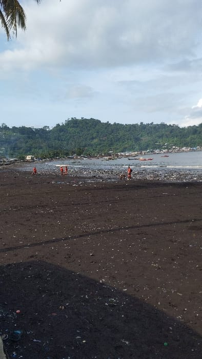 The image shows a black sand beach in Limbe, Cameroon at the coordinates 4.004, 9.211. Several people are walking on the beach, all wearing red shirts. The beach is littered with debris. There is a line of trees on the far side of the beach, followed by water and a series of small boats. The sky is overcast. There are no visible interactions between the people. The image was taken from a distance and appears to be from a smartphone.